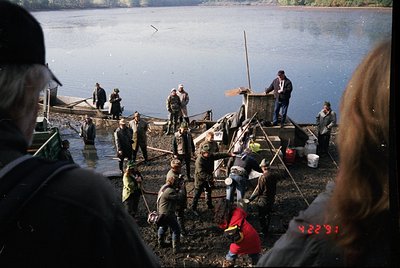 Vintage black-and-white photo of a communal fishing operation on a muddy shore. Men in workwear (overalls, caps) and a few wo...