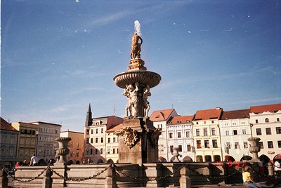 Neoclassical fountain with central statue and cascading water in a European city square, surrounded by multi-story, pastel-co...