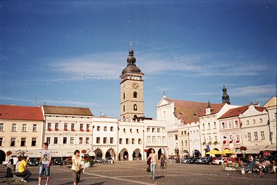 Historic European town square with Baroque-style architecture. Prominent clock tower with spire dominates center, flanked by ...