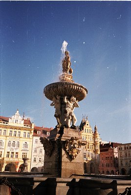 Baroque-style fountain with water spouting from a central urn flanked by sculpted figures, set against historic European arch...