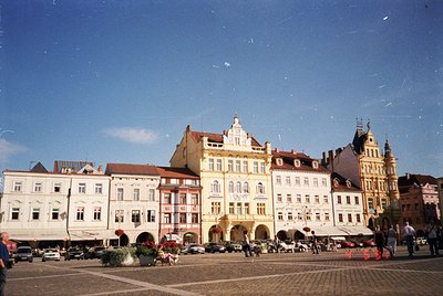 Historic European town square with **Art Nouveau and Baroque** facades. Central building features arched ground-floor entranc...