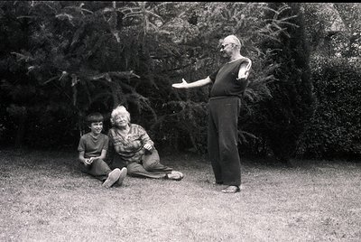 Mid-20th century black-and-white portrait: three individuals in a lush garden setting. A man in a short-sleeve shirt stands g...
