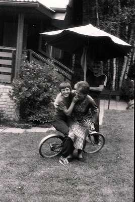 Two children joyfully embrace on a vintage tandem bicycle in a mid-century backyard. The adult in the background under a pati...