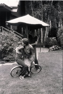 Vintage black-and-white photo of two adults and a child in a lush garden setting, likely mid-20th century. A woman in a strip...