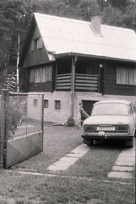 A vintage log cabin-style house with a pitched roof and brick foundation, framed by a simple metal gate. A Soviet-era ZAZ-965...