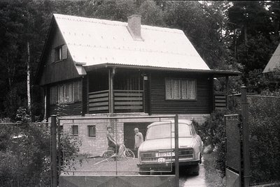 Mid-century wooden house with gabled roof and wooden balcony, framed by chain-link fence. Classic sedan parked beside a child...