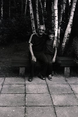 Two generations share a moment in a serene park setting, framed by birch trees. The adult wears a striped shirt and sunglasse...