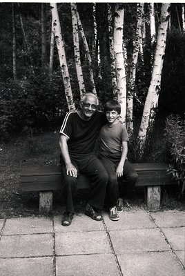 Black-and-white portrait of an adult and child seated on a park bench surrounded by birch trees, likely mid-20th century. The...