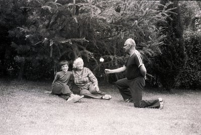 Mid-20th century black-and-white photo: three individuals in a lush garden setting, likely 1950s–1960s. A man kneels, tossing...