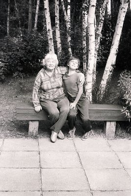 Two individuals sit closely on a wooden bench in a serene park setting, surrounded by birch trees. The elder wears a patterne...