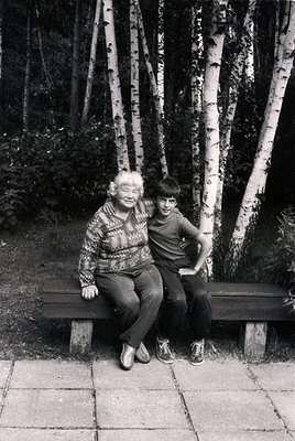 Black-and-white portrait of an elderly woman and young boy sitting closely on a wooden bench in a birch forest. The woman wea...