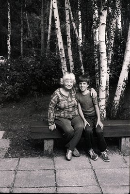 A black-and-white portrait of an elderly woman and young boy seated on a wooden bench in a birch forest. The woman wears a pa...