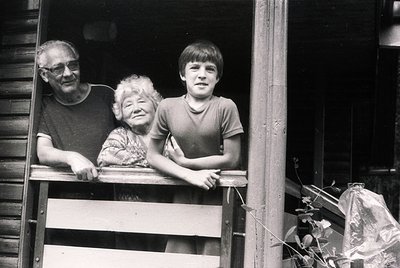 Three generations pose on a wooden balcony, framed by rustic wooden railings. Elderly man and woman in casual 1970s attire (t...