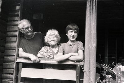 Three-generation family poses on rustic wooden balcony, likely Eastern European rural setting. Elderly man, woman, and young ...