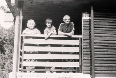 Three generations pose on a wooden porch, framed by rustic railings and a numbered wooden house (). Mid-20th century rural se...