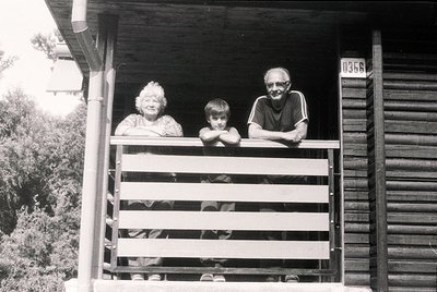 Family portrait on a wooden porch, likely mid-20th century. Three generations pose on a wooden balcony with a rustic wooden h...