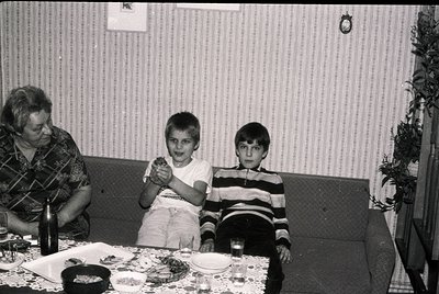 Vintage black-and-white family portrait from the 1970s–80s, featuring a woman and two boys seated at a table with plates of f...