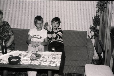 Black-and-white snapshot of two boys seated at a floral-patterned table in a domestic setting, likely mid-1970s. One boy hold...