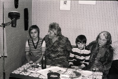 Four generations gathered around a table in a modest indoor setting, likely a 1970s Eastern European home. A woman in pattern...