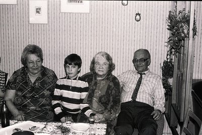 Four generations seated indoors, likely mid-20th century. Elderly woman (left) in patterned sweater, boy in striped shirt, ol...
