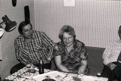Vintage black-and-white photo of three adults seated indoors, likely mid-1970s Eastern Europe. Woman in patterned blouse exam...