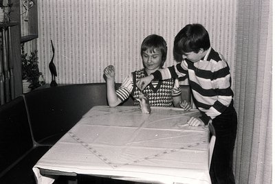 Two children play at a table in a mid-century indoor setting. Girl in patterned sweater leans forward, boy in striped shirt p...