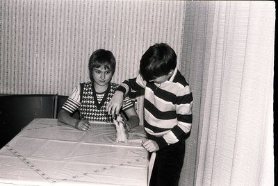 Two boys in 1970s-style striped and patterned sweaters play with a toy train on a tablecloth with floral embroidery. Indoor s...