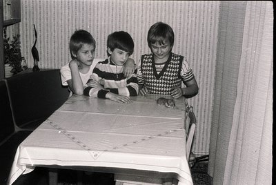 Three children seated at a table in a mid-century indoor setting, likely a 1970s Eastern European home. The boy on the left w...