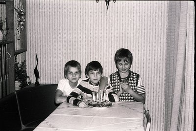 Vintage black-and-white indoor birthday scene: three children pose at a table with a lit cake featuring six candles. Mid-cent...