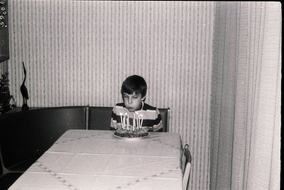 Black-and-white indoor shot of a child seated at a table with a birthday cake adorned with 7 lit candles. Mid-20th century ho...