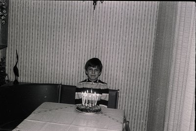 Vintage black-and-white indoor portrait of a young boy seated at a table, blowing out lit candles on a round cake. Plain stri...