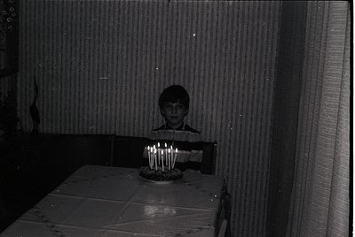 Black-and-white indoor portrait of a child standing beside a table with a lit multi-candle birthday cake. Plain wall and curt...