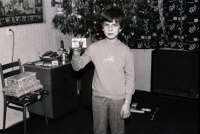Young boy in 1970s-era sweater holds a small electronic device (likely a calculator or toy) in a modestly decorated indoor se...