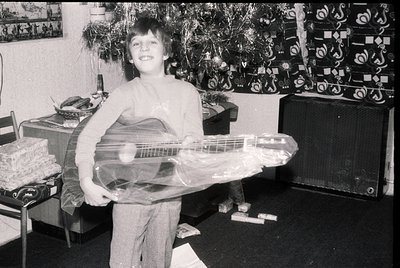 Young boy in 1970s-style sleeveless shirt and trousers poses indoors with a wrapped electric guitar, likely a gift. Surroundi...
