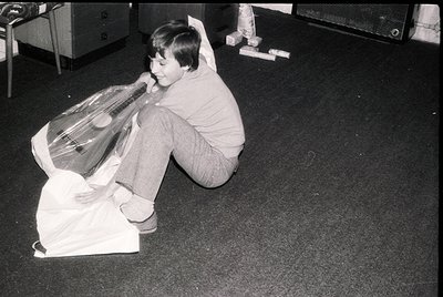 Child playing with a large plastic bag, possibly mimicking a guitar or musical instrument, in an indoor setting. Mid-20th cen...