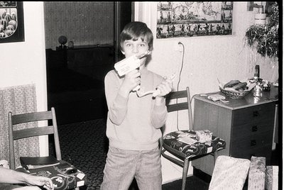 Black-and-white indoor shot of a young boy (mid-1970s) holding a toy gun and a toy sword in a mid-century living room. Wooden...