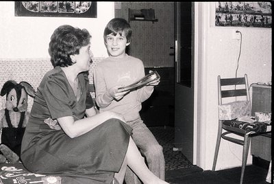 A woman and child in a mid-20th-century living room, likely 1960s–1970s. Woman in a short-sleeve blouse and skirt holds a boo...