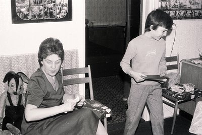 A woman in a 1970s-style blouse works on a sewing project at a wooden table, while a young boy in a long-sleeve shirt and pan...