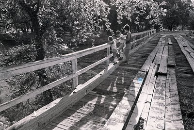 Mid-20th century wooden boardwalk with metal railings, reflecting sunlight on wet planks. Two adults and a child in casual 19...