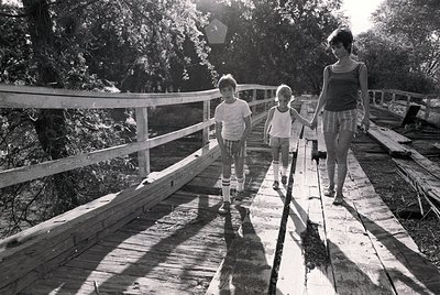 A mid-20th-century black-and-white photo captures a woman and two children walking on a wooden boardwalk through a lush, shad...
