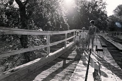 A family of three walks across a weathered wooden boardwalk in a lush, sunlit forest. The adult leads two children, all dress...