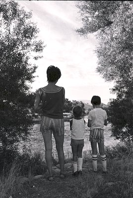 Three figures stand on a rocky riverside in mid-20th century attire, backs to camera. Woman in striped skirt and cardigan, fl...