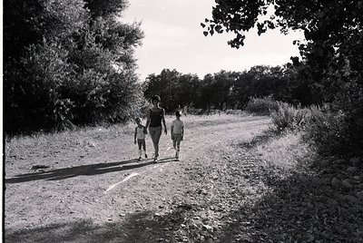 Three figures walk a rural dirt path flanked by dense greenery, captured in monochrome. Adult leads two children, likely mid-...