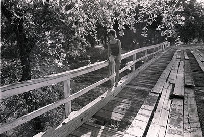 A lone individual stands on a weathered wooden boardwalk, leaning against a metal railing. The scene captures mid-20th centur...
