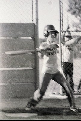 Black-and-white photo of a baseball player mid-swing in a batting cage, wearing a helmet and protective gear. Cage mesh and c...