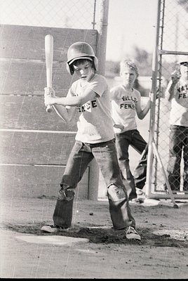 Young baseball player in 1970s-era protective gear—helmet, knee pads, and belt—mid-swing at a school or youth league game. "V...