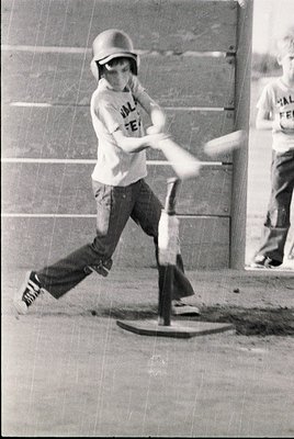 Young baseball player mid-swing in vintage helmet and "Valley Feeder" jersey, 1960s-70s. Indoor concrete court with T-shaped ...