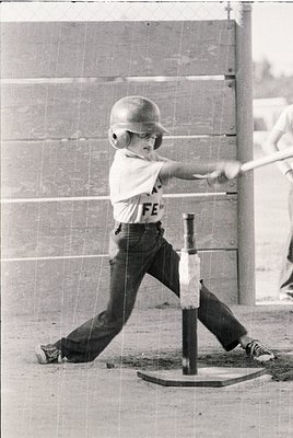 Young baseball player mid-swing in vintage batting helmet and "FE" jersey, 1960s-70s style. Indoor batting cage with wooden f...