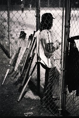 Baseball player in vintage uniform stands at dugout entrance, gripping bat. Chain-link fence and dugout bench visible. Likely...