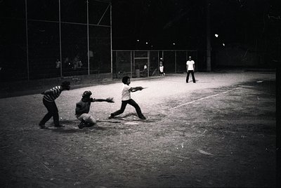 Mid-20th century baseball game under stadium lights, featuring three players in action: batter, catcher, and infielder. Concr...